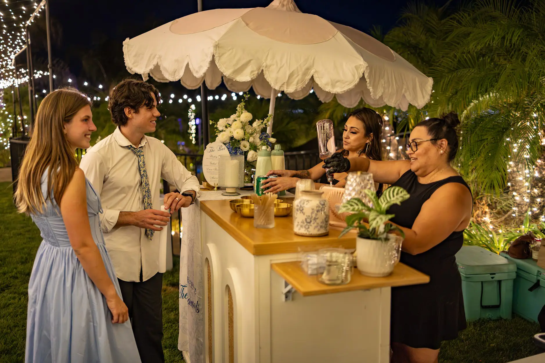 Bartenders in black serving craft sodas from a floral mobile bar at an evening wedding in Redlands